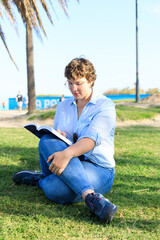 young, overweight woman reading a book on the grass on a sunny day with an urban backdrop