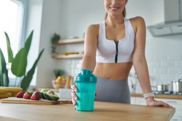 Close-up of woman in sports clothing holding shaker with protein cocktail at the kitchen