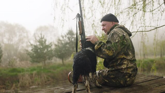 A hunter with a weapon is sitting near a misty lake. His friend, a hunting dog, runs up to him, the dog licks his face, showing his love, which makes his hunting master smile.
