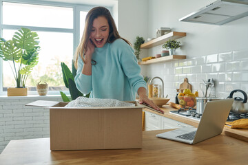 Surprised young woman unpacking box while standing at the domestic kitchen - obrazy, fototapety, plakaty