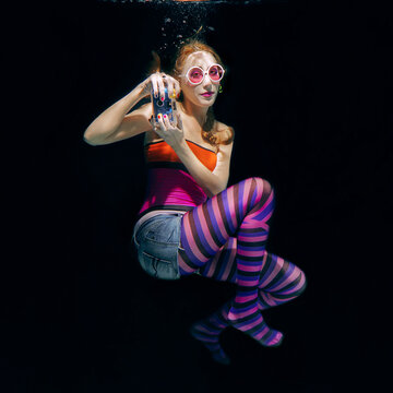 redhead funny woman in colorful clothes and pink sunglasses on the dark background swimming underwater with photo camera. Travel, sightseeing concept