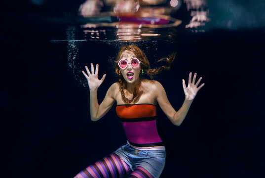 redhead funny woman in colorful clothes and pink sunglasses on the dark background swimming underwater