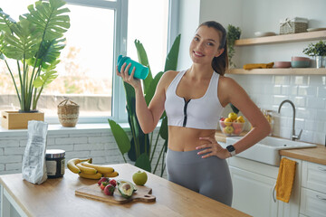 Cheerful young woman in sports clothing preparing protein cocktail at home