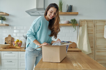 Beautiful young woman unpacking box while standing at the domestic kitchen - obrazy, fototapety, plakaty