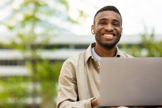 Smiling black man sitting outdoors in park wearing headphones and typing on laptop keyboard while looking at camera