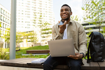 African American guy talking holding smartphone near mouth using voice assistant while sitting on bench with laptop