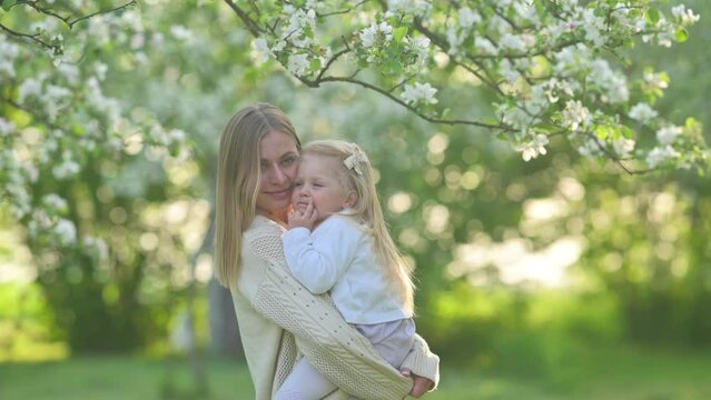 A young cheerful mum is holding a baby in her arms in a beautiful blooming garden while on a summer holiday. Family outdoor lifestyle.