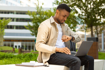 african american man looking at smart watch, sitting on bench with laptop, smart watch