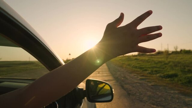 Young woman driver drives car, catches wind with her hand from car window. Free girl in front seat of car, sticking her hand out window, catching wind, glare of sun. Family, child traveling by car