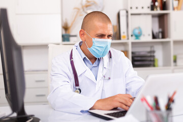 Man doctor in medical mask is working at laptop in clinic office