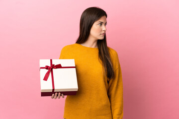 Teenager Brazilian girl holding a gift over isolated pink background looking to the side