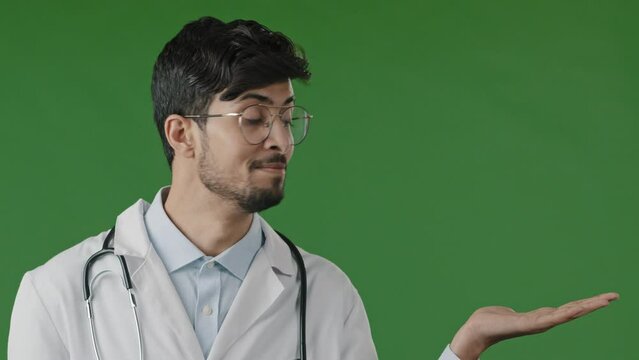 Arab indian guy young male doctor practitioner man hold copy space on palm pointing at workspace mockup promo place for advertisement image offer imaginary object stand isolated over green background