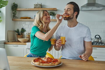 Happy young couple feeding each other with pizza and smiling while sitting at the kitchen island
