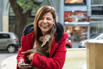 Latin woman laughing while holding her dog and smartphone