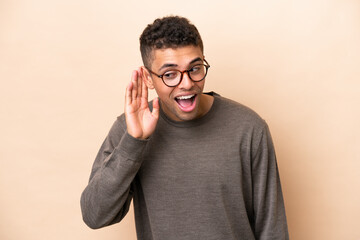 Young Brazilian man isolated on beige background listening to something by putting hand on the ear