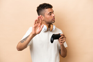 Naklejka na ściany i meble Young Brazilian man playing with a video game controller isolated on beige background making stop gesture and disappointed