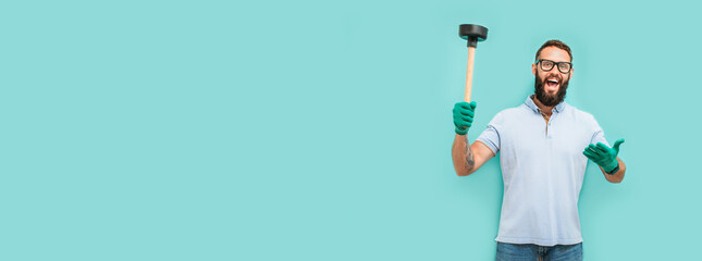 Young handsome man wearing plumber uniform holding toilet plunger looks happy. Professional cleaning of clogged pipes. Studio shot on blue background. Funny promotion poster