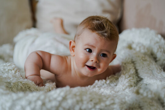  adorable naked 6 month old baby boy lying on belly and looking up in prone position after bathing wrapped in white towel at home on the couch in the living room