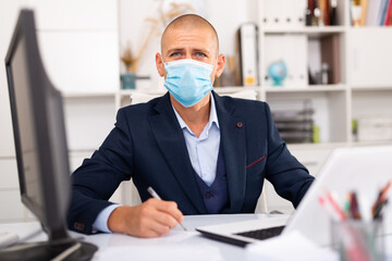 Fototapeta na wymiar Portrait of young man in medical mask sitting at desk with laptop and working in office