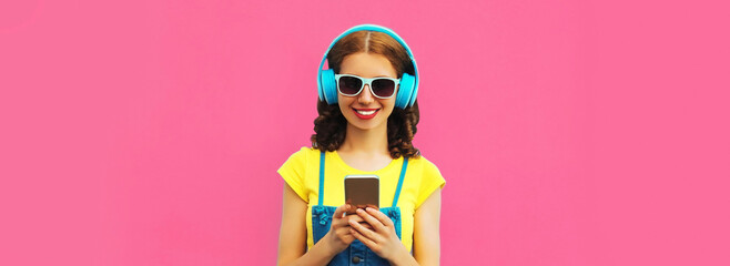 Portrait of smiling young woman in headphones listening to music with smartphone on pink...