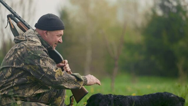 Hunter squatted after a morning hunt with a gun in his hands on a background of the field. His friend the hunting dog runs up to him, the hunter strokes his head with his hand