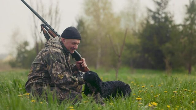 Hunter squatted after a morning hunt with a gun in his hands on the background of the field. He calls a hunting dog. the hunter strokes the dog's hand on his head, in response the dog licks his face