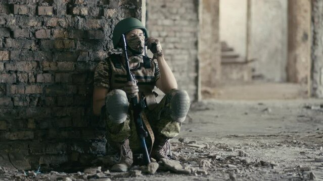 A young military Ukrainian soldier sits on the territory of an abandoned building during the fighting. An explosion occurs and dust falls from the destroyed walls. The concept of the war in Ukraine.