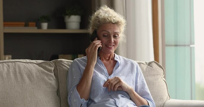 Blond middle-aged woman make call lead conversation on smartphone sit on cozy sofa in living room at home. Older gen using modern tech for receiving remote consultation, personal communication concept