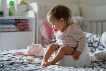 One small caucasian baby girl one year old sitting on the children's potty at home on the bed in...