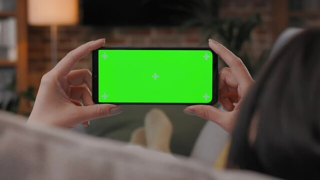 Over shoulder view of young woman holding smartphone with green mock-up screen horizontal mode. Woman lying on couch at home in evening, watching content videos blogs, news or films.Cinematic lighting