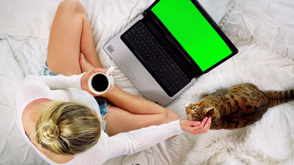Top-down view of a woman sitting on the bed looking at a blank green screen of a laptop computer...