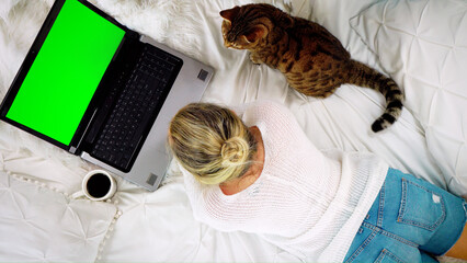Top-down view of a woman lying on the bed looking at a blank green screen of a laptop computer drinking coffee, Bengal cat sitting nearby
