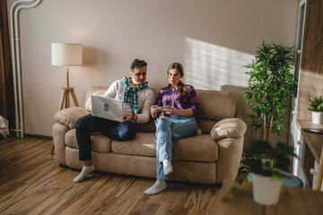 Two people man and woman young adult caucasian couple boyfriend and girlfriend or married husband and wife sitting on the sofa bed at home using mobile phone and laptop for work or leisure