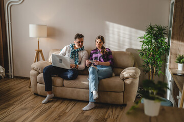 Obraz premium Two people man and woman young adult caucasian couple boyfriend and girlfriend or married husband and wife sitting on the sofa bed at home using mobile phone and laptop for work or leisure