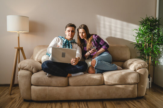 Two people man and woman young adult caucasian couple boyfriend and girlfriend or married husband and wife sitting on the sofa bed at home using mobile phone and laptop for work or internet browsing