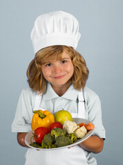 Child chef cook hold plate with vegetables prepares food on isolated grey studio background. Kids cooking. Teen boy with apron and chef hat preparing a healthy vegetables meal in the kitchen.