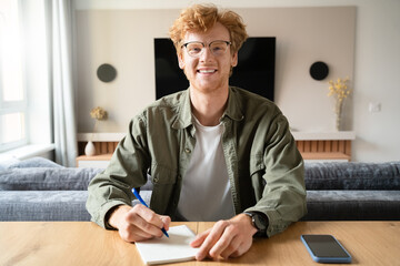 Young man writing notes looking at camera holding videoconference webcam view