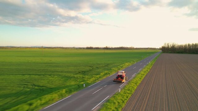 AERIAL: Red tractor with attached farming equipment driving on countryside road. Working farmer returning home with tractor from maintaining agricultural fields. Modern machinery for heavy farm work.