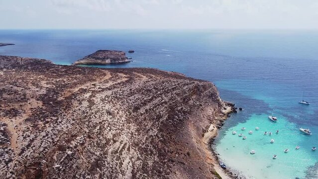 Aerial drone. Cala Pulcino (caletta barche volanti) and Spiaggia and Isola dei Conigli, Lampedusa. Tranquil, cove-style beach with white sand and turquoise surf bordered by rocky cliffs.	