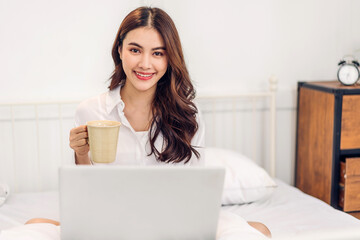 Young smiling happy beautiful asian woman relaxing using laptop computer in the bedroom at home.Young creative girl working and typing on keyboard.work from home concept