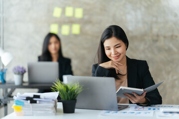 Fototapeta premium Attractive asian young confident businesswoman sitting at the office table with group of colleagues in the background, working on laptop computer.