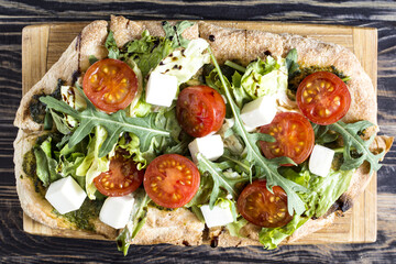 Pizza with cheese, tomatoes, cabbage and arugula on a wooden stand on a natural wooden background.