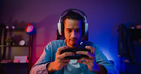 Fotobehang Gaming Happy asia man gamer wear headphone competition play video game online with smartphone colorful neon light in living room at night modern house. Esport streaming game online, Home quarantine activity.  © tirachard