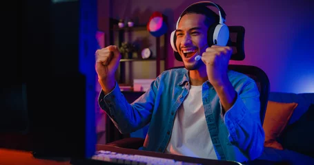 Fotobehang Gaming Happy asia guy gamer wear headphone participation play video game colorful neon lights computer in living room at night modern house. Esport streaming game online, Home quarantine activity concept.  © tirachard