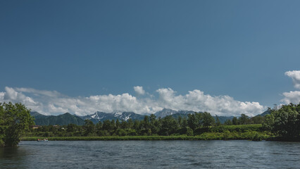 Fototapeta premium A blue calm river flows in the valley. A rafting boat with people is visible. Green vegetation on the shore. Picturesque mountains against the sky and clouds. Copy space. Kamchatka