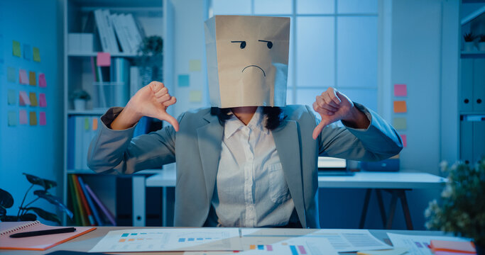 Young Asian Businesswoman Sit On Desk Wear Draw Sad Mask Paper Craft Bag Feel Bad Mood With Fail Work Project In Office At Night. Anonymous Lady Mad And Confuse With Overwork, Work Mental Health.