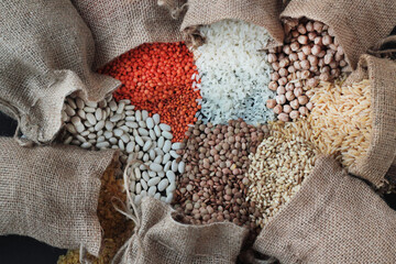 Red lentils, chickpeas, white rice and bulgur pouring from jute sack onto black surface with copy...