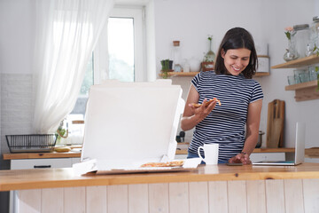 A pregnant woman in the kitchen in her house. A woman drinks tea, looks at her smartphone, and eats pizza. Pizza delivery concept. With a place to copy. High quality photo