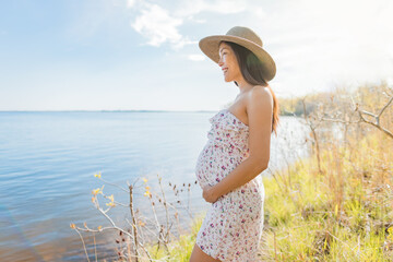 Pregnant Asian woman wearing sun hat during spring nature walk by the lake relaxing enjoying view of natural water landscape. Happy pregnancy outdoors active lifestyle