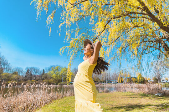 Happy Asian pregnant woman dancing free in nature summer park wearing sun hat and yellow dress. Pregnancy joy and carefree